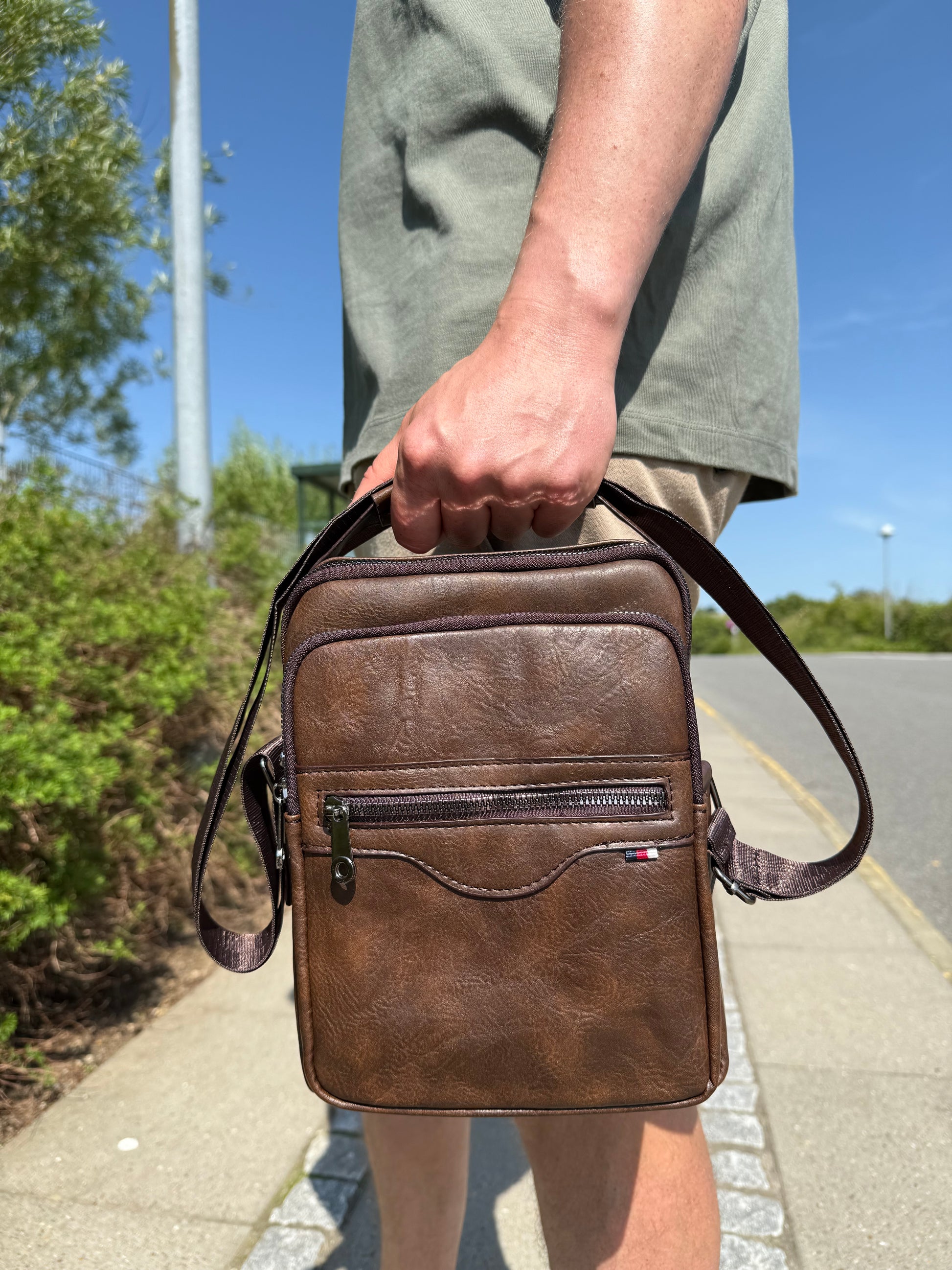 Mandlig model holder Unisex Messenger Taske i mørkebrun i hånden.